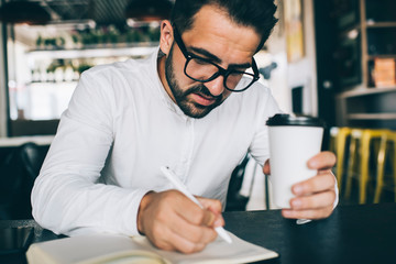 Concentrated man in optical spectacles spending time for planning productive strategy with personal textbook for memo notes, serious male entrepreneur with coffee to go writing checklist in notepad