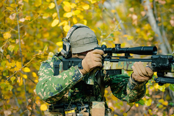 Airsoft man in uniform with sniper rifle, make correction in optical sight on yellow forest...