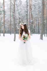 Winter wedding. Beautiful young bride in the winter snowy forest, wearing elegant wedding dress, holding bouquet and wreath on the head.