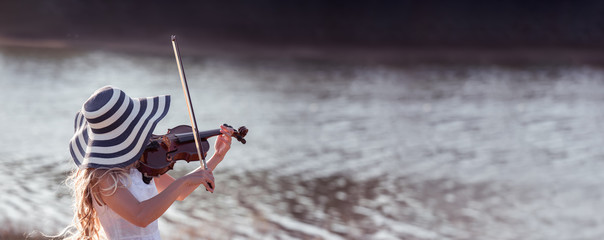 Back view of woman with hat raised arms holding violin at the river.