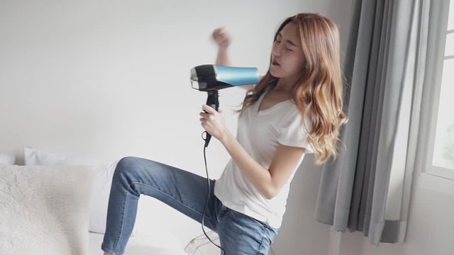 Funny asian kid singing into hairdryer and crazily jumping on bed, dreaming of becoming famous artist. She couldn't stop herself swaying. She can't calm down.