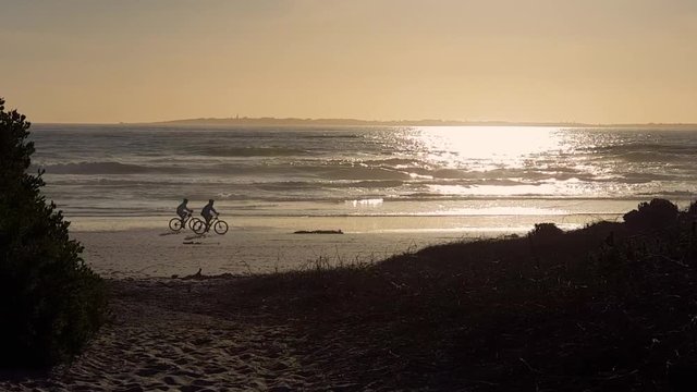Silhouettes of cyclists riding on wet shiny beach sand, on holiday fun. Waves peacefully breaking. Sun glistening on flat ocean. Robben Island visible on horizon. Footprints in sand path to beach.
