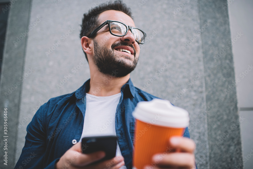 Poster below view of millennial bearded male blogger with takeaway cup in hand looking away and smiling dur