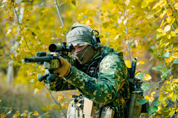 Airsoft man in uniform, stand with sniper rifle on yellow forest backdrop. Soldier aims at the sight