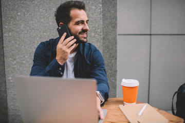 Fototapeta na wymiar Happy male freelancer enjoying break for smartphone calling to best friend using roaming connection outdoors, cheerful bearded man making international conversation while working remotely in cafe