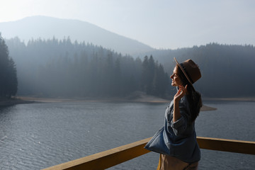 happy young woman in hat enjoys lake view in mountains . Relaxing moments in forest. Back view of...
