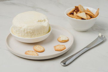 Cheese and cracker in a white bowl with a knife on a white table.