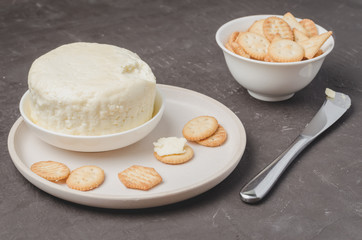 Cheese and cracker in a white bowl with a knife on a dark stone background.