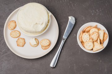 Cheese and cracker in a white bowl with a knife on a dark stone background. Top view.