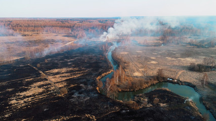 Black burned field, a bush burns in the distance and there is a strong thick smoke. A small river blocks the path of fire.