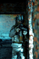 Fototapeta na wymiar Man in uniform with machine gun and night-vision device stand beside broken wall. Airsoft soldier in night building. Vertcal photo