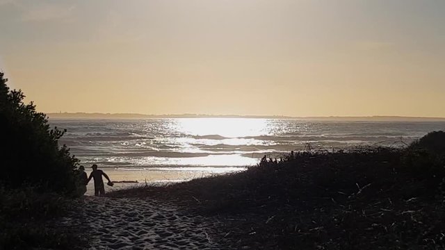 Silhouettes of romantic couple in love. Walking hand in hand over sea sand to beach. Mirror shiny wet sea sand. Sunlight glistening on ocean. Golden waves break on beach. Robben Island on horizon.