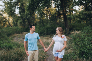 Naklejka na ściany i meble Portrait of a young family expecting a baby. A loving man and a beautiful pregnant blond woman holding hands at sunset in the forest. Pregnancy picture, concept.