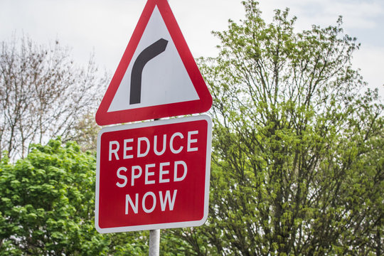 Red and white REDUCE SPEED NOW sign located in the UK with a background of green trees and plants on a public highway.