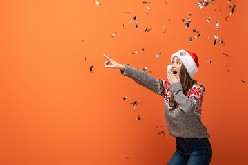 surprised woman in santa hat under falling confetti pointing with finger away on orange background