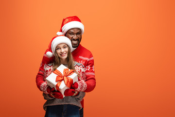 happy interracial couple in santa hats and Christmas sweaters hugging and holding gift on orange background