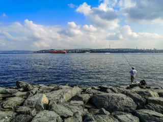 Fototapeta na wymiar Fisherman fishes in the Sea of ​​Marmara on the promenade in Istanbul, Turkey. Large boulders on the banks of the Bosphorus, a large red tanker at sea, and a cityscape on the other side of the bay