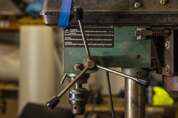Close up of an industrial drilling machine held partly together with blue tape, covered in dust, dirt, shavings, and grime. A three pronged handle on the side of the drill giving this retro machine us