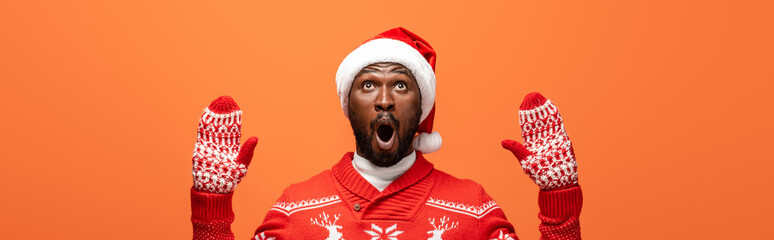 shocked african american man in santa hat, mittens and Christmas sweater isolated on orange, panoramic shot
