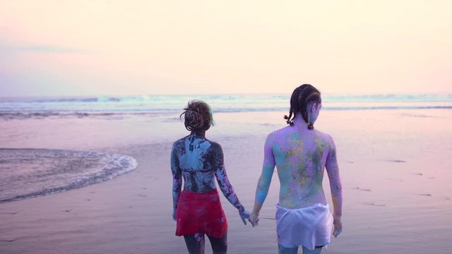 Rear view of a young adult сaucasian couple with painted bodies walk along the beach hand in hand