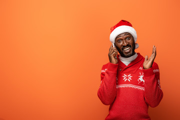 surprised african american man in santa hat and Christmas sweater talking on smartphone on orange background