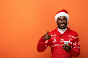 smiling african american man in santa hat and Christmas sweater with smartphone showing thumb up on...