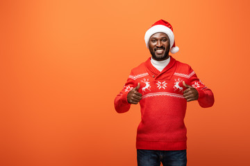 Fototapeta na wymiar smiling african american man in santa hat and Christmas sweater showing thumbs up on orange background