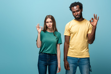 Fototapeta premium displeased interracial couple showing ok sign on blue background