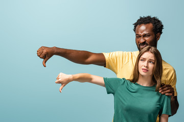 displeased interracial couple showing thumbs down isolated on blue background