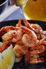 Shrimp fried in a pan with garlic and lemon on a black plate and a glass of beer on a gray background. Hand squeezes a lemon