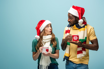 surprised interracial couple in santa hats and scarves holding mugs with cocoa and looking at each other isolated on blue