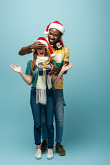 happy african american man in santa hat and scarf gifting present to surprised girlfriend and covering eyes on blue background