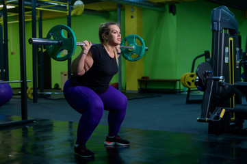 Fototapeta na wymiar Overweight woman crouches with a barbell in the gym. A fat girl does exercises on the buttocks. Goes in for sports for weight loss.