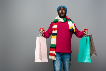 shocked african american man in winter outfit with shopping bags on grey background