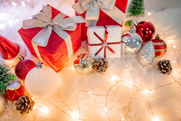Christmas decoration on white hairy carpet with illuminated lights. Gift box with ribbon, glossy ball, pine cone and Christmas tree on fluffy rug. Present and decorative objects for New Year holiday