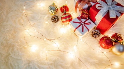 Christmas decoration on white hairy carpet with illuminated lights. Gift box with ribbon, glossy ball and pine cone on fluffy rug. Present and decorative objects for New Year holiday season.