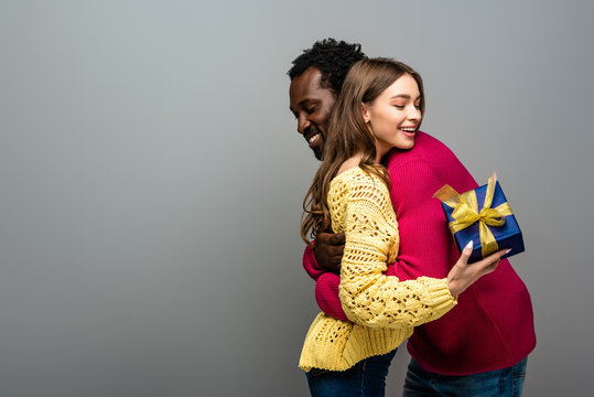happy interracial couple in sweaters holding present and hugging on grey background