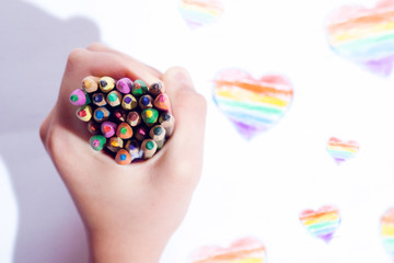 A child's hand holds colored pencils for drawing on a background of painted hearts in the gamma of...