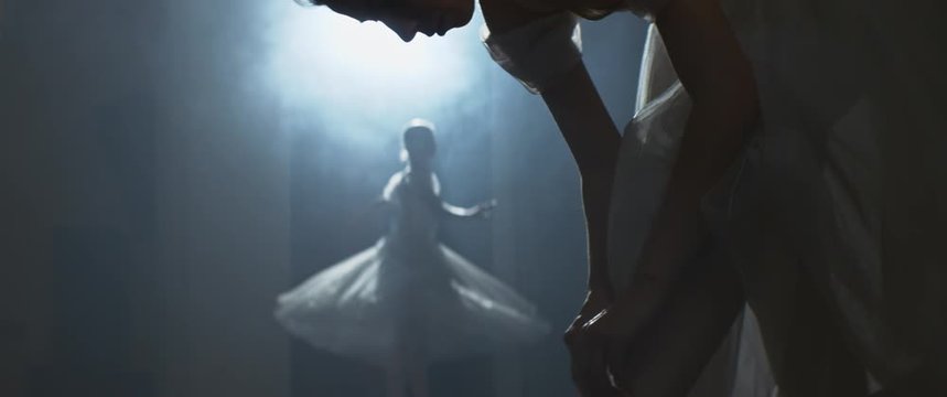 Ballerina touching pointe shoes at backstage of rehearsal or performance. Ballet dancer spinning in the background on stage in costume in spotlight, Anamorphic lens, handheld shot
