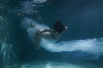 Fototapeta na wymiar Beautiful girl swims underwater with long hair. Blue or gold background like gold. The atmosphere of a fairy tale or magic. Diving under the water with a shiny cloth