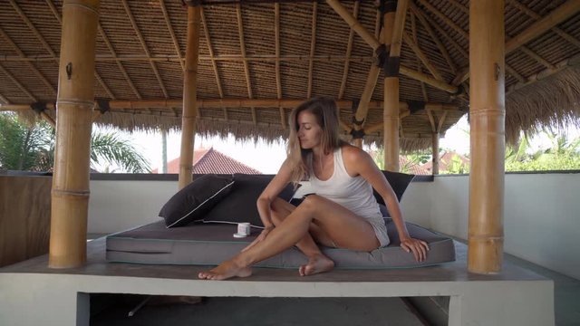 Beautiful young woman sitting on mat in thatched patio, smiling and applying after-sun lotion to skin on her legs after sunbathing in tropics