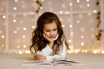 Happy curly little child girl is reading fairy tale book on the background with lights. Merry...