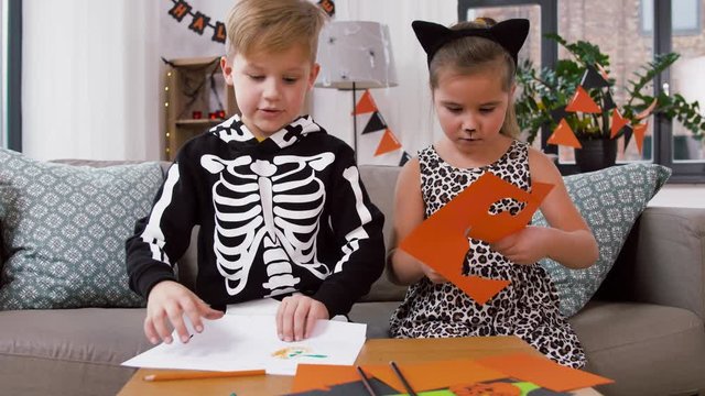 halloween, holiday and childhood concept - little boy and girl in costumes drawing and doing crafts at home