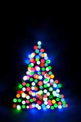 Christmas fairy lights are defocused in the shape of a fir tree, giving a blurry effect. Dark background for design.
