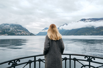 Beautiful caucasian woman stands on pier in Lugano. Girl in travel. Lake Lugano, southern slope of Alps. Landscape in Switzerland. Amazing scenic outdoors view. Canton of Ticino. Adventure lifestyle