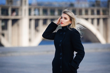 A blonde in a black classic coat walks on unfinished bridges over the river in the fall. Autumn photo shoot in the city