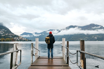 Fototapeta na wymiar Man with backpack enjoy panorama on pier in Lugano. Man in travel. Lake Lugano, southern slope of Alps. Landscape in Switzerland. Amazing scenic outdoors view. Canton of Ticino. Adventure lifestyle