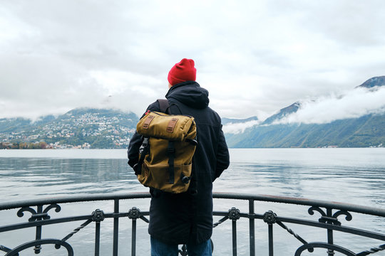 Man With Backpack Enjoy Panorama On Pier In Lugano. Man In Travel. Lake Lugano, Southern Slope Of Alps. Landscape In Switzerland. Amazing Scenic Outdoors View. Canton Of Ticino. Adventure Lifestyle