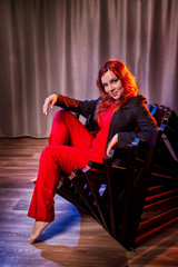 Beautiful young woman with redhair, black hat and red dress on black strange armchair in the room and nice light behind her