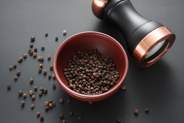 Pepper mill with copper inserts on black background with some black pepper in a clay bowl.